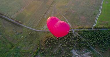 Red aerostat over fields. Romantic hot air balloon in a form of a heart travels in the sky on beautiful nature background at sunset. Top aerial view. video
