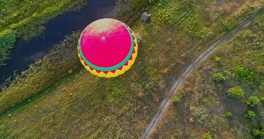 Aerostat on nature background. Colorful hot air balloon flying over the field near the small river. Outdoor activity. Top view. Camera moves down. video