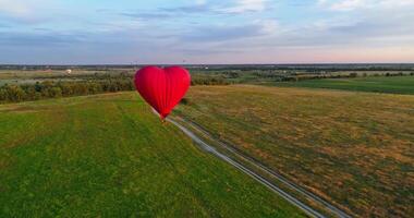 Travel of romantic hot air balloon. Red aerostat balloon flying over the fields under blue sky. Beautiful aerostat in the form of a heart.Aerial view. video
