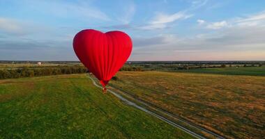 Hot air balloon flying over field. Romantic red aerostat in the heart shape moving slowly in the air. Fantastic view on the airship in summer. video