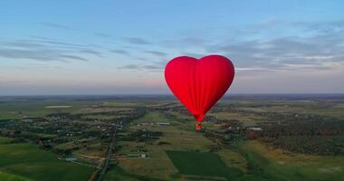 Flight of a red airship. Beautiful hot air balloon in heart shape travelling over the green fields in the morning. Aerostat with basket in the sky. video