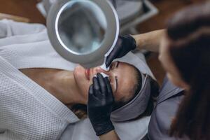 Cosmetologist in rubber gloves performs facial cleansing using a magnifying glass with LED light lupa photo