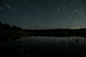 In the dark of night, star trails create a mesmerizing display above a calm lake. The still water reflects the light and surrounding trees, enhancing the tranquil atmosphere. photo