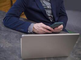 Businessman in blue suit jacket and checkered shirt sitting at desk, multitasking with smartphone and laptop Focused on productivity while effectively managing tasks using laptop in office environment photo