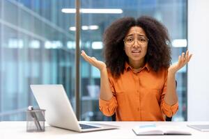 Confused woman in office setting expressing uncertainty. Dressed in orange shirt, sitting at desk with open laptop and book, raising hands in confusion, seeking help. photo