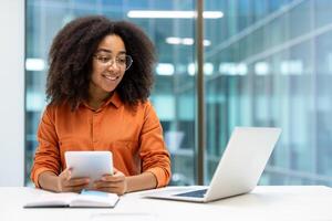 Professional woman engaging with digital technology including a tablet and laptop in an office environment. The modern workspace features glass walls photo