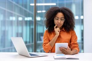Thoughtful professional woman using tablet and laptop in bright office. Focused on digital task, embracing modern technology. Ideal for business, technology, productivity, work concepts photo