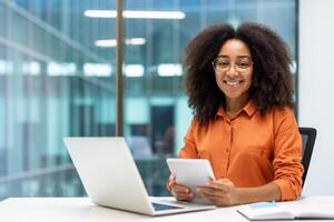 Portrait of young female programmer inside office at workplace, businesswoman smiling and looking at camera, holding tablet computer in hands, testing new software, sitting at table with laptop. photo