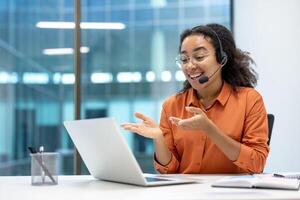 A cheerful businesswoman wearing a headset engages in a conference using her laptop. She gestures expressively while seated in a modern office space, emphasizing connectivity and communication photo