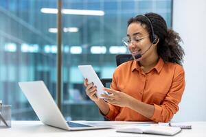 Woman with headset and tablet computer working inside office with laptop, support and helpline worker. photo