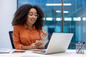 A business professional engages in a virtual meeting using her laptop, jotting down notes in her notebook. She is in a modern office setting with large glass windows photo