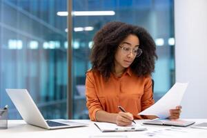 Professional woman working with documents at desk. She reviews information with laptop and papers. This setting conveys business, concentration, and productivity in modern office. photo