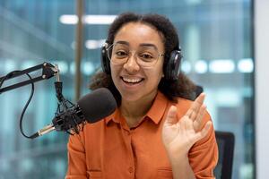 Smiling young woman using microphone and headphones for podcast in studio. She waves confidently, creating warm, engaging atmosphere. Ideal for topics like broadcasting, recording, media, communication. photo
