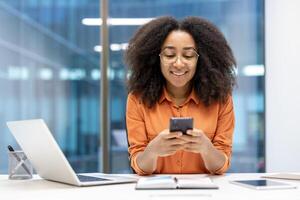 Smiling woman in office uses smartphone while working, multitasking with laptop and tablet. Displays concept of digital communication, productivity, and positive work environment. photo