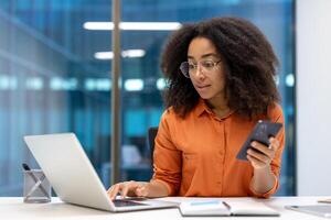 Confident woman working with a laptop and phone in a contemporary office space. She is multitasking by focusing on both devices, showcasing productivity and professionalism. photo