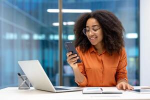 Smiling young woman checking phone at desk with laptop in modern office setting. Displays professional and positive work environment, blending technology and productivity. photo