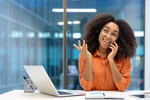 Confident African American businesswoman engaged in phone conversation in bright office. Smiling young professional using smartphone and working on laptop for communication and networking. photo