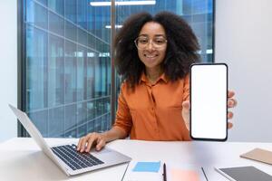 A cheerful individual showcasing a blank smartphone screen while working on a laptop at a bright modern office setting. photo