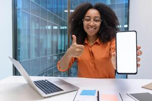 Smiling woman with glasses and afro hair holds smartphone with blank screen and gives thumbs-up gesture, seated at desk with laptop in office. Ideal for concepts of positivity, technology, approval. photo