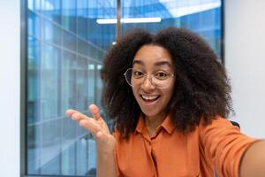 Happy young woman takes selfie with phone in modern office, displaying joy and connection. Bright smile and engaging expression capture positive communication. photo