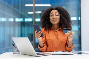 A woman at her desk displaying a smartphone and credit card with a concerned look, suggesting potential financial or technology issues. She is surrounded by a laptop, notepad photo