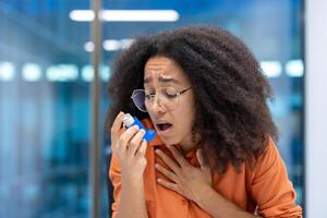 Concerned young woman using asthma inhaler in modern office. She holds her chest, appearing distressed. Office setting visible in background, conveying need for health care support. photo