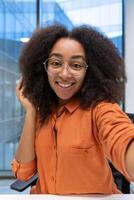 Confident young woman wearing glasses and smiling while taking a selfie at work. The image displays natural lighting, a bright atmosphere, and modern architecture visible through large glass windows photo
