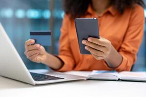 Young African American businesswoman using phone and credit card for online shopping. Engaging in digital transactions at work with laptop, showcasing modern professional lifestyle. photo