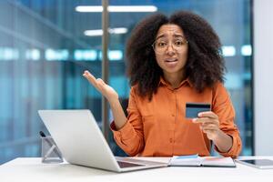 A woman in a bright office looks at the camera with concern, holding up a credit card working at her laptop. She appears perplexed, suggesting an issue with online payments or banking. photo