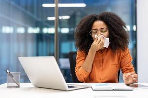 Woman in office sneezing at desk using tissue, illustrating cold symptoms and health awareness. Concept of work during illness and need for self-care in professional setting. photo