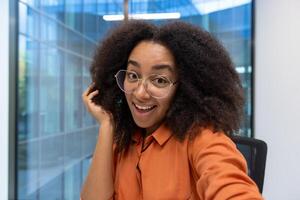 Young woman at workplace smiling and looking into phone camera, businesswoman talking happily with colleagues using call app on smartphone. photo