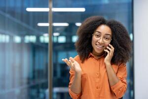 Portrait of a joyful businesswoman, office worker with a phone in her hands talking to clients, smiling and looking at the camera, standing near the window of an office building at her workplace. photo