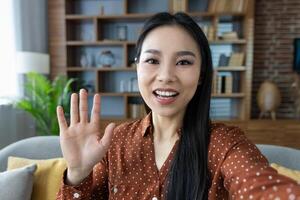 Asian woman smiling while waving to camera, engaging in friendly online call at home, showcasing joy and connection. Great for virtual communication imagery. Modern, cozy setting. photo