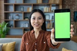 Asian woman in casual attire shows smartphone with vibrant green screen at home. Focus on phone that is held towards camera, inviting concepts of technology, communication, apps. photo