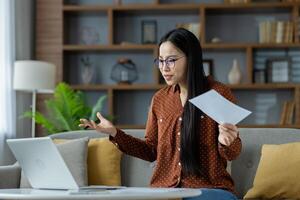 Asian woman in home office setting, engaged in call holding paper, discussing project. Laptop on table, remote work environment. Emblematic of modern communication and indoor workspace. photo