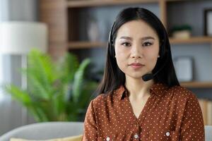 Asian woman wearing headset sitting in home setting, engaged in online communication. Ideal representation of remote work, tech interaction, customer support. Calm, professional demeanor. photo