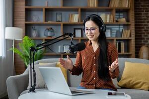 Asian woman hosting a podcast at home, speaking into a microphone while using a laptop. Wearing headphones, she appears engaged and lively, surrounded by a comfortable setting. Smartphone visible. photo