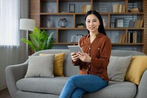 Portrait of young Asian woman, student smiling and looking at camera holding tablet computer, studying remotely sitting at home on sofa in living room, using online app. photo