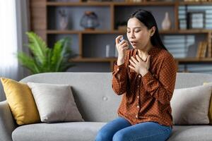 Asian woman using asthma inhaler, sitting on sofa experiencing breathing issue. Wearing polka dot blouse, managing respiratory health at home. Focused, calming atmosphere. photo