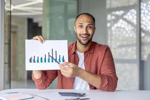 Successful man with document, graph, report looking at camera, showing colleagues and partners successful results of achievement, using meeting, online call, working inside office. photo