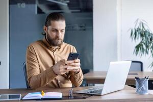 A professional dressed in smart casual attire focuses on his smartphone while seated at an office desk, working alongside a laptop and a notepad. photo