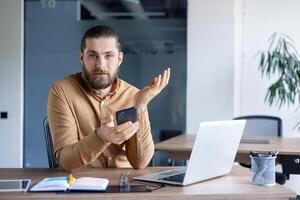 Professional office setting with a man holding a smartphone while working on a laptop. Modern workspace showing casual workwear and productivity tools like notebooks and writing utensils. photo