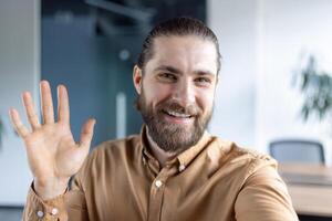 Friendly bearded professional making a warm gesture during a webcam meeting in a modern office setting. photo