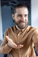 A confident young man with a welcoming smile and a long beard gestures warmly in a bright office setting. He seems approachable and cheerful, embodying positivity and friendliness. photo