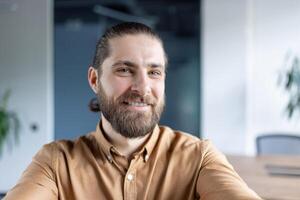 Happy young man with a beard taking a selfie in a contemporary office setting. Showcasing a friendly attitude, casual style, and a professional interior environment. photo