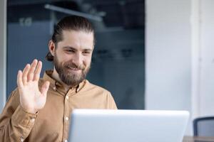 A bearded man in a casual shirt participates in a call, waving and smiling warmly at the screen. Ideal representation for communication, remote work, or online professional interactions. photo