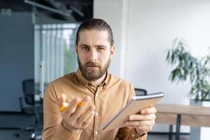 Focused professional man holding a notebook, looking judgmental, inquisitive, and serious, likely during a business interaction in an office environment photo