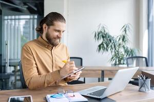 Smiling businessman working at a desk, writing in a notebook with a laptop nearby. Ideal for concepts of productivity, business planning, and professional work environments. photo