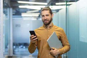 Young professional dressed in business casual attire holds a laptop and smartphone while standing in a modern office environment, showcasing confidence and tech-savviness. photo