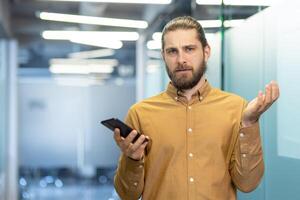Man standing in a modern office environment holding a smartphone, expressing confusion. Professional attire and demeanor indicate a workplace-related situation. photo